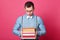 Indoor studio shot of dark haired confused model posing isolated over bright pink background, holding books in his strong hands,