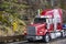 Industrial grade powerful red big rig semi truck with flat bed semi trailer running with rain dust on the wet road with protective