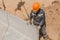 An industrial worker in overalls and a protective helmet attaches a heavy iron hook lifting mechanism with a steel cable rope to a