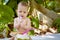 Infant baby at tropical vacation. Eats and drinks green young coconut. Sits on a ground and gnaw a spoon. Jungles on background.