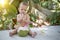 Infant baby at tropical vacation. Eats and drinks green young coconut. Sits on a ground and hold a spoon. Jungles on background