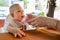 Infant girl in white dress sitting at the table on a high chair. Baby drink water from the bottle from mother hands on the street