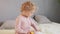 Infant wavy haired blond girl sitting in bed eating dried apricots with pleasure and feeding her toy having fun wearing ping