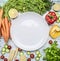 Ingredients for cooking salad cherry tomatoes, lettuce, peppers, spices and oil laid out around white plate on wooden rustic bac