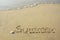 The inscription `summer` by handwriting and shell on the beach sand with the soft wave and sunlight in the evening