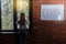 The inscription on the wall is the word COVID. A young woman is standing near a window in a room with a brick wall