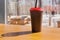 Inside the forest cafe, there is coffee in a black paper cup on a wooden table. The park is visible outside the window