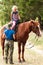 The instructor helps the girl to saddle a brown horse in the forest. Children`s equestrian camp. Summer sports camp for children.