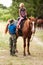 The instructor helps the girl to saddle a brown horse in the forest. Children`s equestrian camp. Summer sports camp for children.