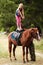 The instructor helps the girl to saddle a brown horse in the forest. Children`s equestrian camp. Summer sports camp for children.