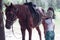 The instructor helps the girl to saddle a brown horse in the forest. Children`s equestrian camp. Summer sports camp for children.