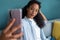 Interesting young african american woman using her mobile phone to take a selfie while sitting on sofa at home