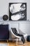 Interior design of living room with stylish navy blue commode, grey armchair, pillow, black clock, mock up modern paintings.