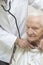 An internist doctor examines the lungs of a very old gray-haired woman sitting in a chair with a stethoscope.