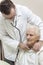 An internist doctor examines the lungs of a very old gray-haired woman sitting in a chair with a stethoscope.