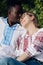 Interracial happy couple sits on bench in garden dressed in Ukrainian embroidered shirts