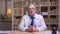 Interview of senior doctor in white coat talking into camera on book shelves background.