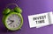 INVEST TIME in an office on a notepad purple background with a green table clock