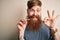 Irish redhead man with beard holding dental invisible aligner for tooth correction doing ok sign with fingers, excellent symbol