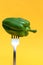 An iron fork is stuck in a green bell pepper, on a yellow background.