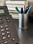 Iron tumbler with pens and pencils, landline black phone is on office desk with books in blurred focus.