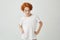 Isolated portrait of funny ginger boy in white t-shirt holding hands on waist, looking in camera with confident