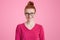 Isolated shot of happy ginger woman in pink sweater, wears square spectacles, has red hair tied in knot, freckled skin, pleasant s