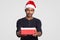 Isolated shot of serious black man wears Santa Claus headgear, carries little box of present, dressed in black jumper, poses over