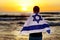 Israeli patriot with the flag of Israel on his shoulders stands on the shores of the Mediterranean Sea at sunset, Israel