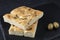 Italian food - focaccia bread with rosemary and olive oil and two green olives on a serving board - black background, side view