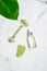 Jade roller with natural anti aging jade stone, essential oil dropper bottle and green leaf on marble table. Facial skin care