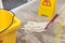 A janitor cleans the surface of a wet concrete floor of a factory with a string mop. A wet floor sign warns passersby