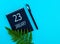 January 23th. Day 23 of month, Calendar date. Black notepad sheet, pen, fern twig, on a blue background