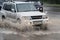 Japanese automobile Mitsubishi Pajero driving flooded street road over deep muddy puddle, splashing drop spray water from wheels