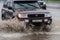 Japanese car Toyota Land Cruiser driving on flooded city street road over deep muddy puddle, splashing drop of spray water from wh