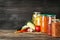 Jars with pickled products and fresh vegetables on wooden table against black background