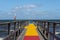 A jetty with a colourful carpet in blue, yellow and red. A blue ocean and sky in the background