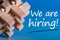 Job recruiting advertisement represented by `WE ARE HIRING` texts on blue background with wooden brain teaser talking