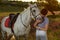 Jockey young girl petting and hugging white horse in evening sunset