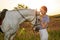 Jockey young girl petting and hugging white horse in evening sunset. Sun flare