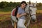 Jockey young girl petting and hugging white horse in evening sunset. Sun flare