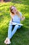 Jocund female student showing thumbs up studying in park and sitting on green grass.