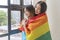 Joy romantic LGBT asian lesbian woman embracing girlfriend from back standing together beside window with rainbow flag at home.