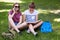 Joyful adorable female tenager speaks via smart phone sits near her nerd friend who reads book, pose together on green grass, enjo