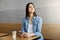Joyful attractive girl with dark hair sitting in cafe, drinks coffee and chatting with friend on smartphone then turning