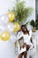 Joyful, attractive, smiling multiracial woman in white dress sit in armchair with golden, silver balloons at home party