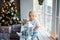 Joyful baby boy in white t-shirt with bear sitting on window with white lantern in hands and looking away in christmas time at