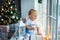 Joyful baby boy in white t-shirt with bear sitting on window with white lantern and looking away in christmas time at home.