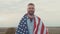 Joyful bearded American man with an American flag on shoulders smiles as looks into the camera and stands in the steppe.