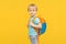 Joyful boy child stands with a backpack on his back and looks at the camera on a bright yellow background